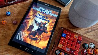 The AD&D Player's Handbook on a tablet sitting on a wooden table, surrounded by a dice box, other books, and a plant pot